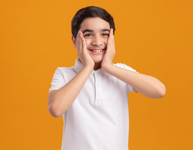 Fröhlicher junger kaukasischer Junge, der Hände auf das Gesicht legt und gerade isoliert auf orangefarbener Wand mit Kopierraum schaut