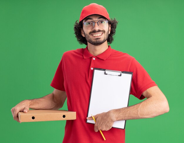 Fröhlicher junger kaukasischer Lieferbote in roter Uniform und Mütze mit Brille, die Pizzapaket mit Bleistift hält und Klemmbrett zur Kamera zeigt