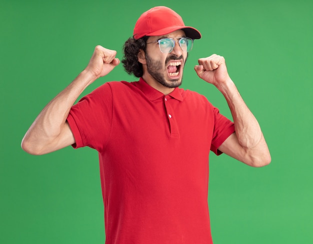 Fröhlicher junger Liefermann in roter Uniform und Mütze mit Brille, der nach vorne schaut und Ja-Geste einzeln auf grüner Wand macht