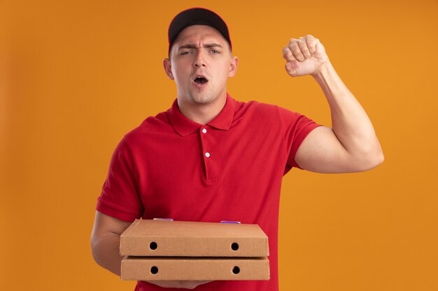 Fröhlicher junger Liefermann in Uniform mit Mütze mit Pizzakartons, die Ja-Geste einzeln auf oranger Wand zeigt