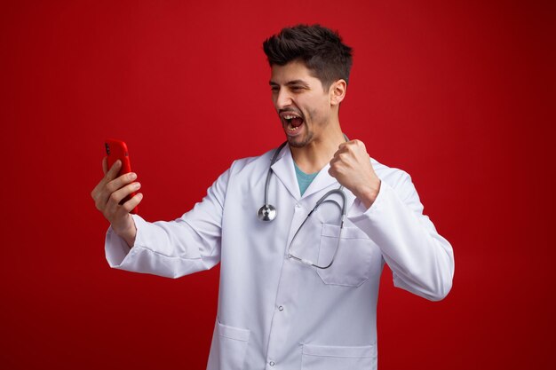 Fröhlicher junger männlicher Arzt, der eine medizinische Uniform und ein Stethoskop um den Hals trägt und das Handy hält und betrachtet, das eine Ja-Geste zeigt, die auf rotem Hintergrund isoliert ist