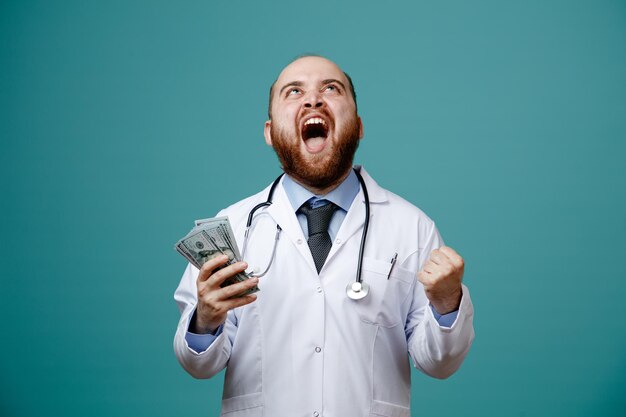 Fröhlicher junger männlicher Arzt mit Arztkittel und Stethoskop um den Hals, der Geld hält und nach oben schaut und Ja-Geste isoliert auf blauem Hintergrund zeigt
