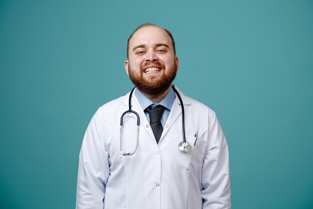 Fröhlicher junger männlicher Arzt mit Arztkittel und Stethoskop um den Hals, der isoliert auf blauem Hintergrund in die Kamera blickt