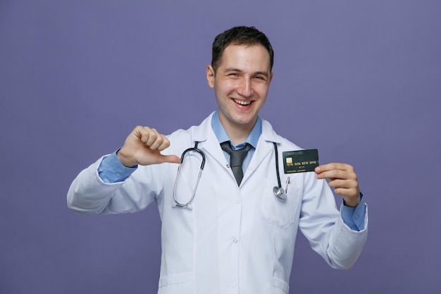 Fröhlicher junger männlicher Arzt mit medizinischem Gewand und Stethoskop um den Hals, der die Kreditkarte zeigt, die darauf zeigt und die Kamera isoliert auf violettem Hintergrund betrachtet