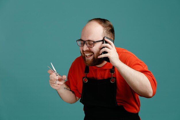 Kostenloses Foto fröhlicher junger männlicher friseur mit brille, rotem hemd und friseurschürze, der eine schere hält und mit geschlossenen augen lacht, während er telefoniert, isoliert auf blauem hintergrund