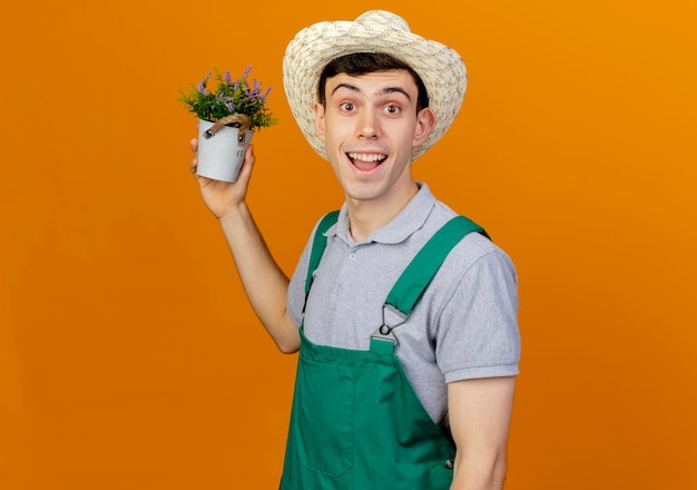 Fröhlicher junger männlicher Gärtner mit Gartenhut hält Blumen im Blumentopf