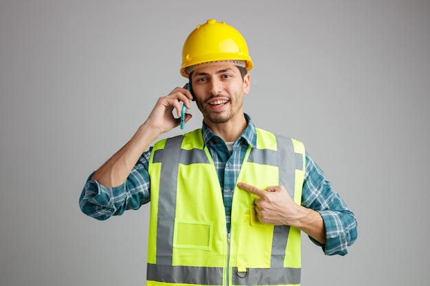 Fröhlicher junger männlicher Ingenieur mit Schutzhelm und Uniform, der in die Kamera blickt, die auf sich selbst zeigt, während er am Telefon isoliert auf weißem Hintergrund spricht