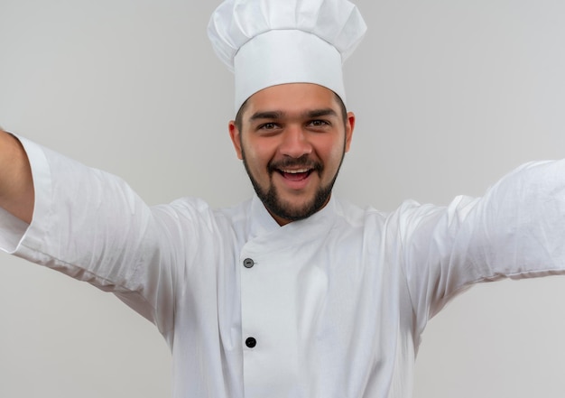 Fröhlicher junger männlicher Koch in Kochuniform, der mit offenen Armen isoliert auf weißer Wand schaut