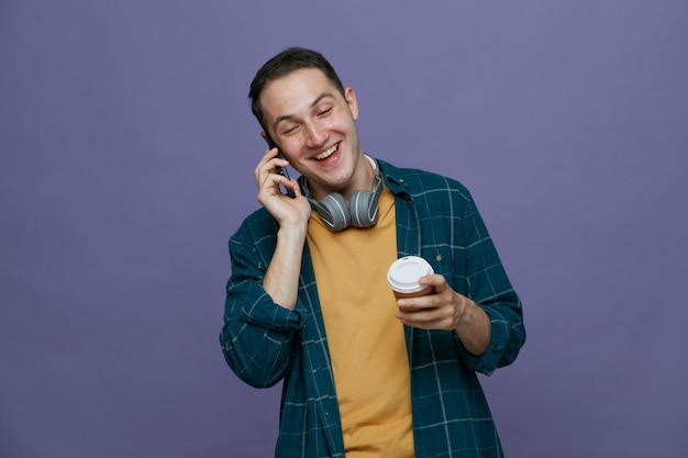 Fröhlicher junger männlicher Student, der Kopfhörer um den Hals trägt und eine Kaffeetasse aus Papier hält, die lachend nach unten schaut, während er am Telefon isoliert auf lila Hintergrund spricht
