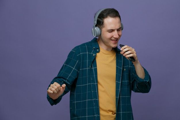 Fröhlicher junger männlicher Student mit Kopfhörern, der ein Handy in der Nähe des Mundes hält und Musik hört, die mit dem Handy als Mikrofon tanzt, gibt vor, mit geschlossenen Augen zu singen, isoliert auf violettem Hintergrund