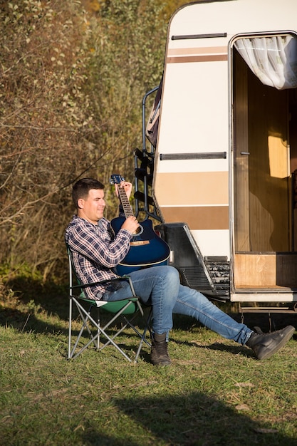 Fröhlicher junger Mann, der auf seiner Gitarre vor seinem Retro-Wohnmobil in den Bergen spielt. Mann, der sich in den Bergen entspannt.