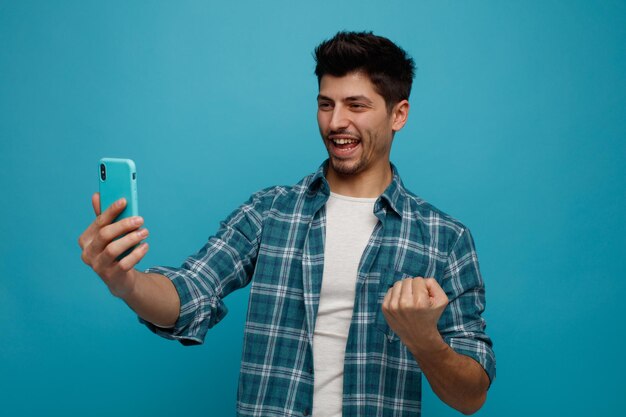 Fröhlicher junger Mann, der das Handy ausstreckt und es anschaut, zeigt Ja-Geste isoliert auf blauem Hintergrund