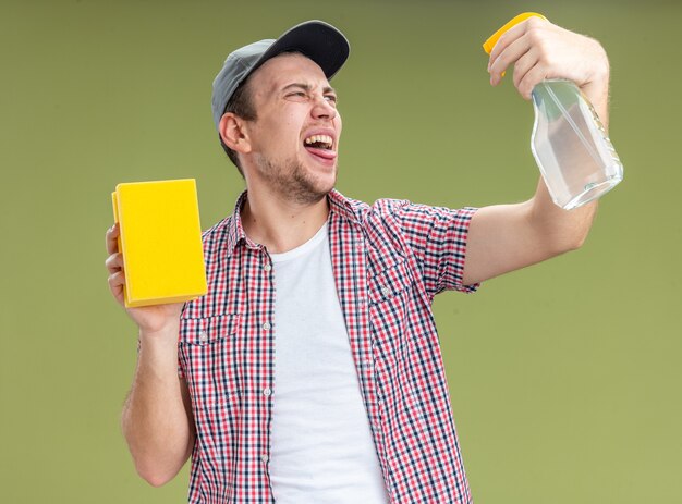 Fröhlicher junger Mann, der eine Mütze trägt und einen Schwamm hält, der sich mit Reinigungsmittel isoliert auf olivgrüner Wand wässert?