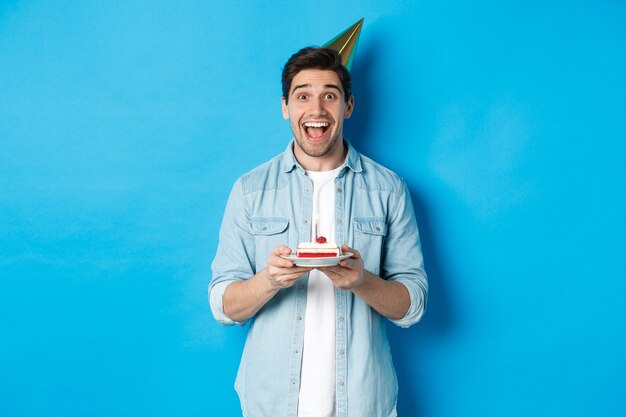 Fröhlicher junger Mann, der Geburtstag im Partyhut feiert, B-Day-Kuchen hält und vor blauem Hintergrund steht.