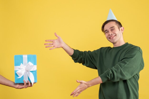 Fröhlicher junger Mann der Vorderansicht, der auf das Geschenk in der menschlichen Hand auf Gelb zeigt