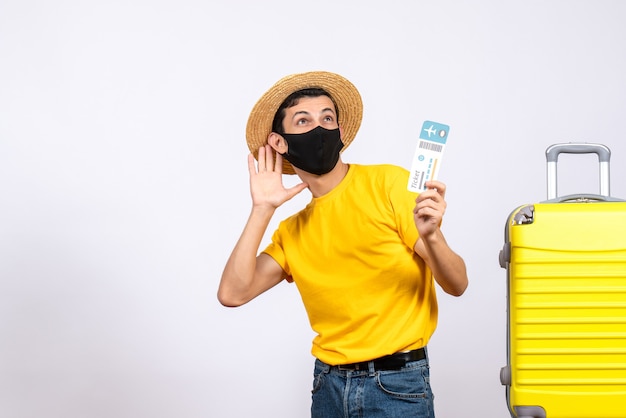 Fröhlicher junger Mann der Vorderansicht im gelben T-Shirt, der nahe gelbem Koffer steht, der Reiseticket hält