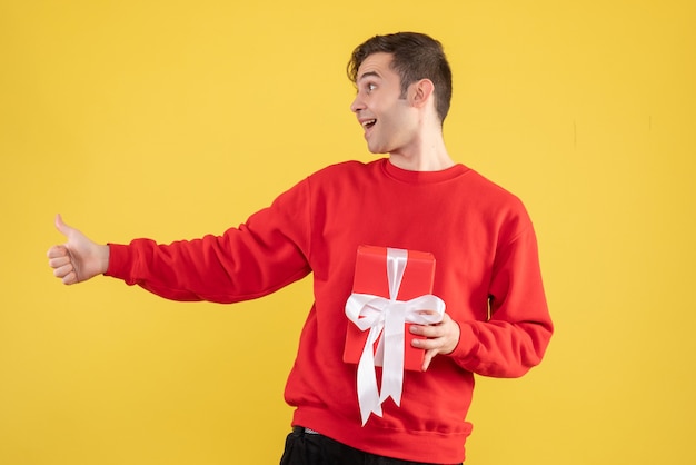 Fröhlicher junger Mann der Vorderansicht mit dem roten Pullover, der Daumen auf Zeichen gelb macht