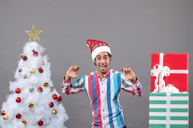 Fröhlicher junger Mann der Vorderansicht mit geöffneten Händen, die nahe weißem Weihnachtsbaum stehen