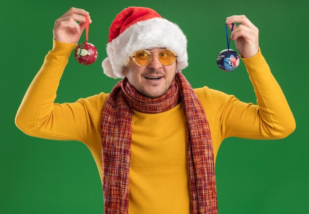 Fröhlicher junger Mann in gelbem Rollkragenpullover und Weihnachtsmütze mit Brille, der Spielzeug für den Weihnachtsbaum hält und in die Kamera schaut, die fröhlich über grünem Hintergrund lächelt