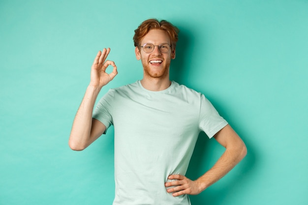 Fröhlicher junger Mann mit roten Haaren und Bart, Brille und T-Shirt, zufrieden lächelnd und mit OK-Zeichen, Ja sagen, genehmigen und zustimmen, über türkisfarbenem Hintergrund stehend