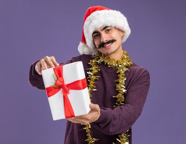 Fröhlicher junger Mann mit Schnurrbart, der Weihnachtsmütze mit Lametta um den Hals trägt und Weihnachtsgeschenk mit einem Lächeln auf dem Gesicht über der lila Wand zeigt