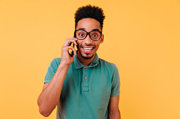 Fröhlicher Kerl im trendigen grünen T-Shirt, der am Telefon spricht. Sorgloser junger Mann, der Smartphone hält und lächelt.