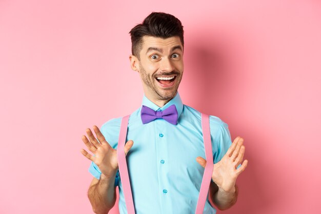 Fröhlicher Kerl in Fliege, der seine Hosenträger zeigt und glücklich lächelt, sich optimistisch fühlt, aufgeregt über rosafarbenem Hintergrund steht.