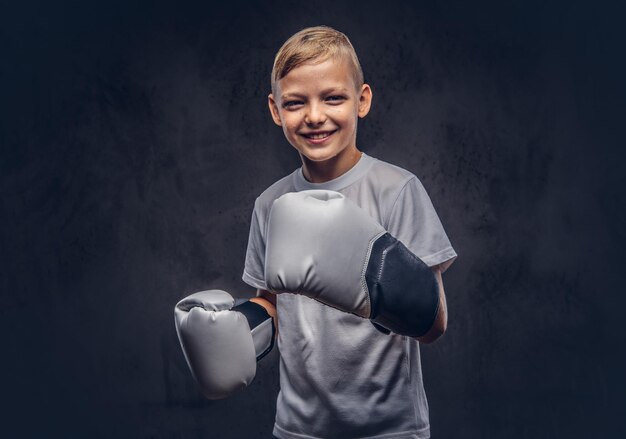 Fröhlicher kleiner Boxer mit blonden Haaren, gekleidet in ein weißes T-Shirt mit Boxhandschuhen, die in einem Studio posieren. Getrennt auf dem dunklen strukturierten Hintergrund.