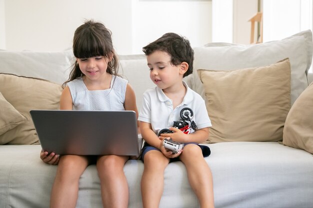 Fröhlicher kleiner Bruder und Schwester, die zu Hause auf der Couch sitzen, Laptop benutzen, Video, Zeichentrickfilme oder lustigen Film ansehen.