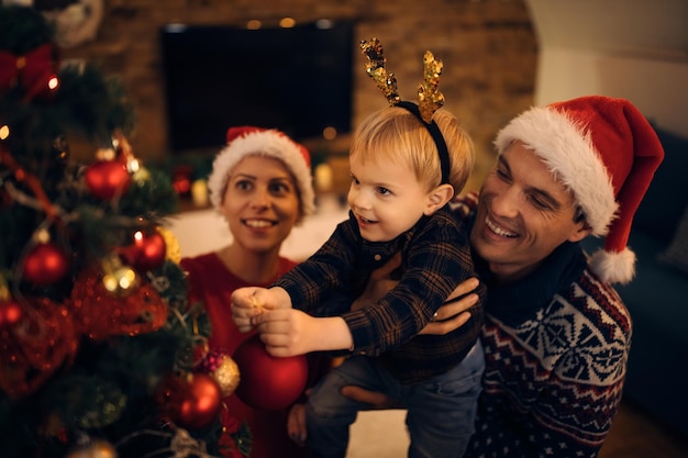 Fröhlicher kleiner Junge und seine Eltern schmücken den Weihnachtsbaum zu Hause