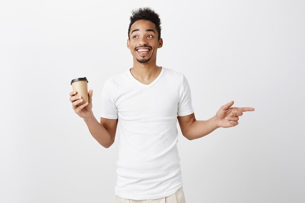 Fröhlicher lächelnder Afroamerikaner Kerl im weißen T-Shirt, das rechts zeigt und Tasse mit Kaffee hält