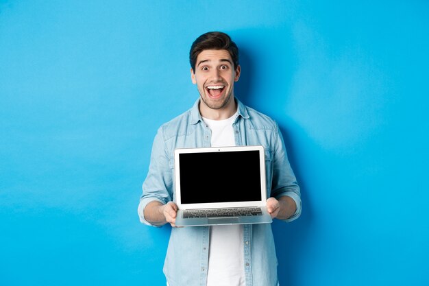Fröhlicher lächelnder Mann, der eine Präsentation macht, einen Laptop-Bildschirm zeigt und glücklich aussieht und auf blauem Hintergrund steht