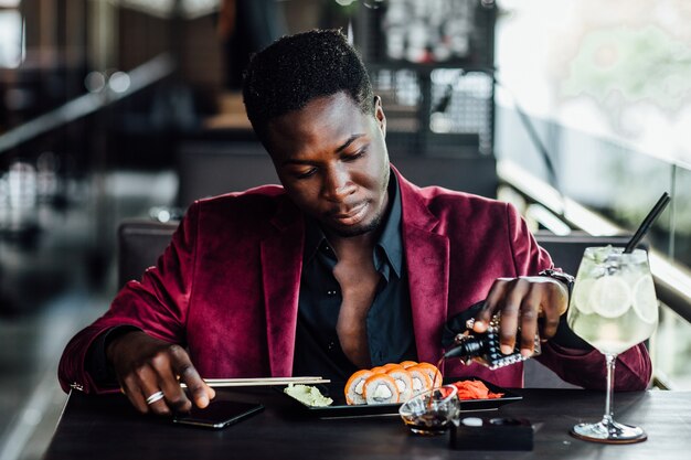 Fröhlicher, lockiger Afrikaner, der Essstäbchen-Sushi-Rollen hält. Chinesische Fischrestaurantterrasse.