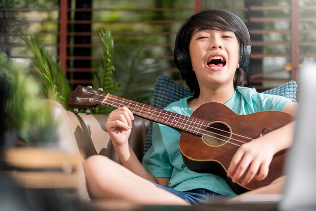 Fröhlicher, lustiger asiatischer Junge, der lautes Spiel Ukulele mit aufregendem und energiegeladenem asiatischen männlichen Sohn singt, macht lautes, lustiges Geräusch, das Ukulele-Online-Unterricht zu Hause übt, zu Hause bleiben Konzept