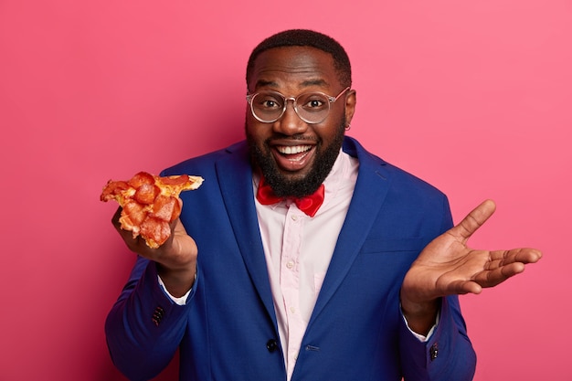 Fröhlicher männlicher Chef hat nach der Arbeit einen Snack, hält appetitliche Pizza, formell gekleidet, hebt die Handfläche, trägt eine Brille, isst Fast Food