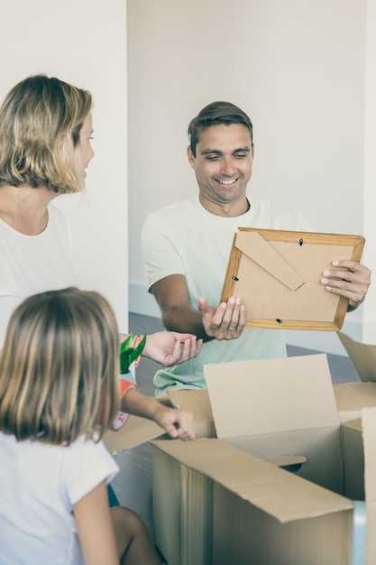 Fröhlicher Mann, der Dinge mit seiner Frau und seinen Kindern in der neuen Wohnung auspackt und auf dem Boden in der Nähe von offenen Kisten sitzt