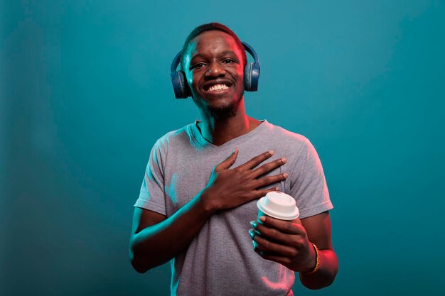 Fröhlicher Mann, der eine Tasse Kaffee hält und Musik auf dem Headset genießt und ein offenes Lächeln vor der Kamera zeigt. Zuversichtliches Modell, das drahtlose Kopfhörer verwendet, um Aufnahmelieder zu hören, während er ein Getränk in der Hand hat.