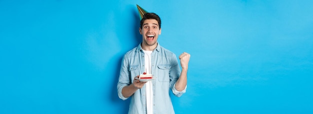 Fröhlicher Mann, der glücklich aussieht, Geburtstag in Partyhut zu feiern, Geburtstagskuchen hält und Faustpumpe st macht