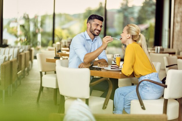 Fröhlicher Mann füttert seine Freundin mit einem Kuchen in einem Café