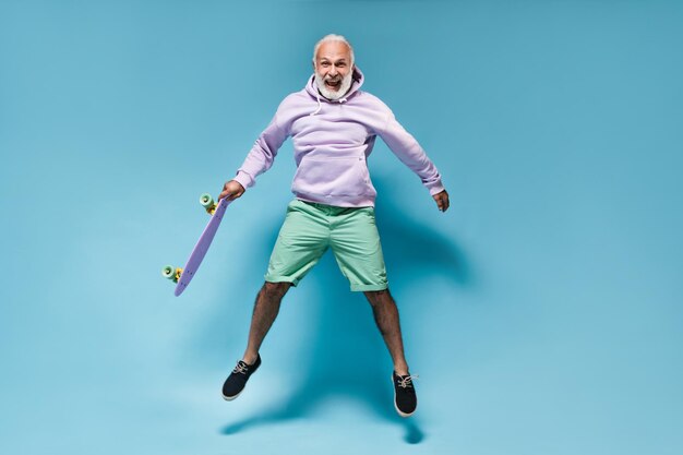 Fröhlicher Mann im Hoodie, der mit Skateboard auf blauem Hintergrund springt Fröhlicher bärtiger Kerl in lila Kleidung und Hemdspringen