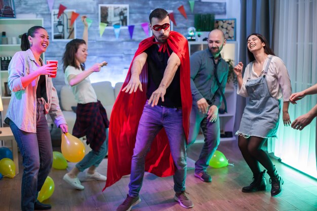 Fröhlicher Mann im Superheldenkostüm, der seine Tanzbewegungen auf der Party von Freunden zeigt.