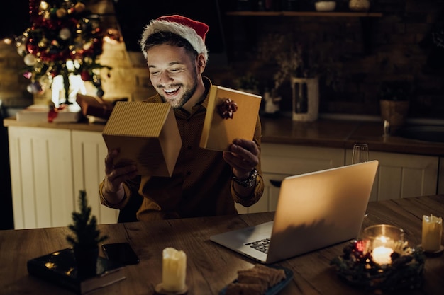 Fröhlicher Mann öffnet Geschenkbox, während er an Heiligabend Videoanrufe über Laptop tätigt