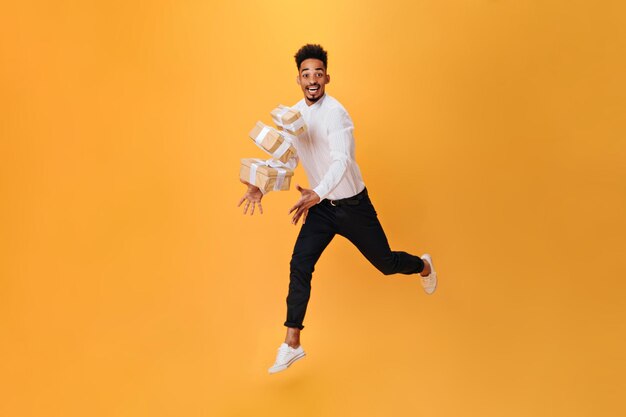 Fröhlicher Mann springt auf orangefarbenen Hintergrund und wirft Geschenke hoch Dunkelhaariger Mann in weißem Hemd und schwarzer Hose posiert glücklich isoliert
