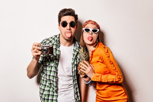 Fröhlicher Mann und Frau in der Sonnenbrille zeigen Zungen und posieren mit Retro-Kamera und Ananas auf isoliertem Raum.