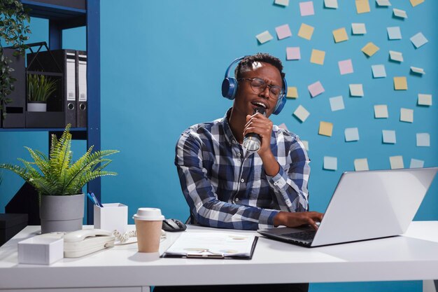 Fröhlicher Mitarbeiter im Büro des Marketingunternehmens, der moderne drahtlose Kopfhörer trägt, während er Musik hört und singt. Glücklicher positiver Geschäftsmann, der am Schreibtisch im Arbeitsbereich sitzt und dabei das Lied genießt.