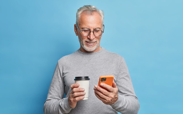 Fröhlicher moderner älterer Mann verwendet Handy für Kommunikationstypen Textnachricht auf Telefonbildschirm hält Pappbecher Kaffee rollt Internetseiten, gekleidet in Freizeitkleidung, isoliert über blauer Wand