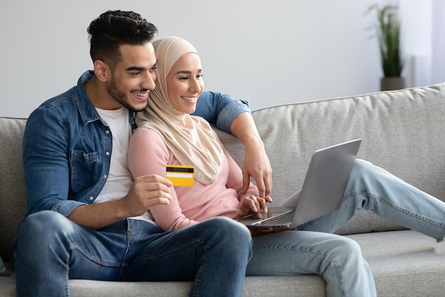 Fröhlicher nahöstlicher familieneinkauf im internet von zu hause aus, mit laptop und kreditkarte, panorama mit kopierraum. arabischer mann und frau im hijab, die online für waren und dienstleistungen bezahlen