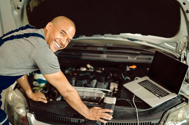 Kostenloses Foto fröhlicher schwarzer mechaniker, der in der autowerkstatt an der motordiagnose arbeitet und in die kamera schaut