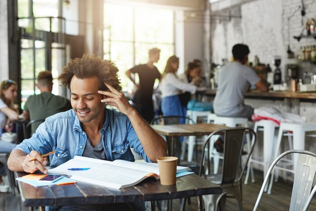 Fröhlicher schwarzer Student mit stilvollem Afro-Haarschnitt, der breit liest, während er Nachricht auf Handy liest, Internet während der Mittagspause surft, während Hausaufgaben im Café macht