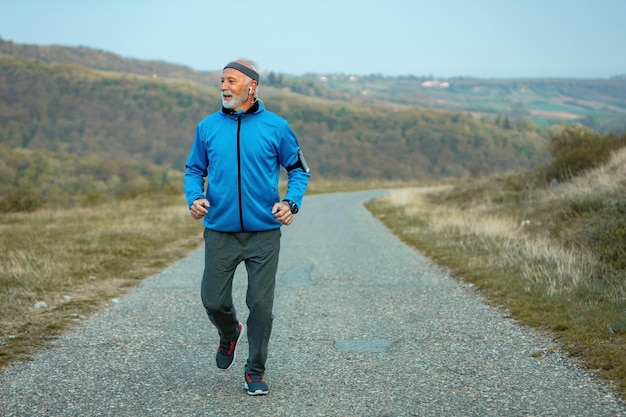 Fröhlicher Senior-Sportler, der sich entschlossen fühlt, mit dem aktiven Lebensstil Schritt zu halten und auf der Straße in der Natur zu laufen Kopierraum